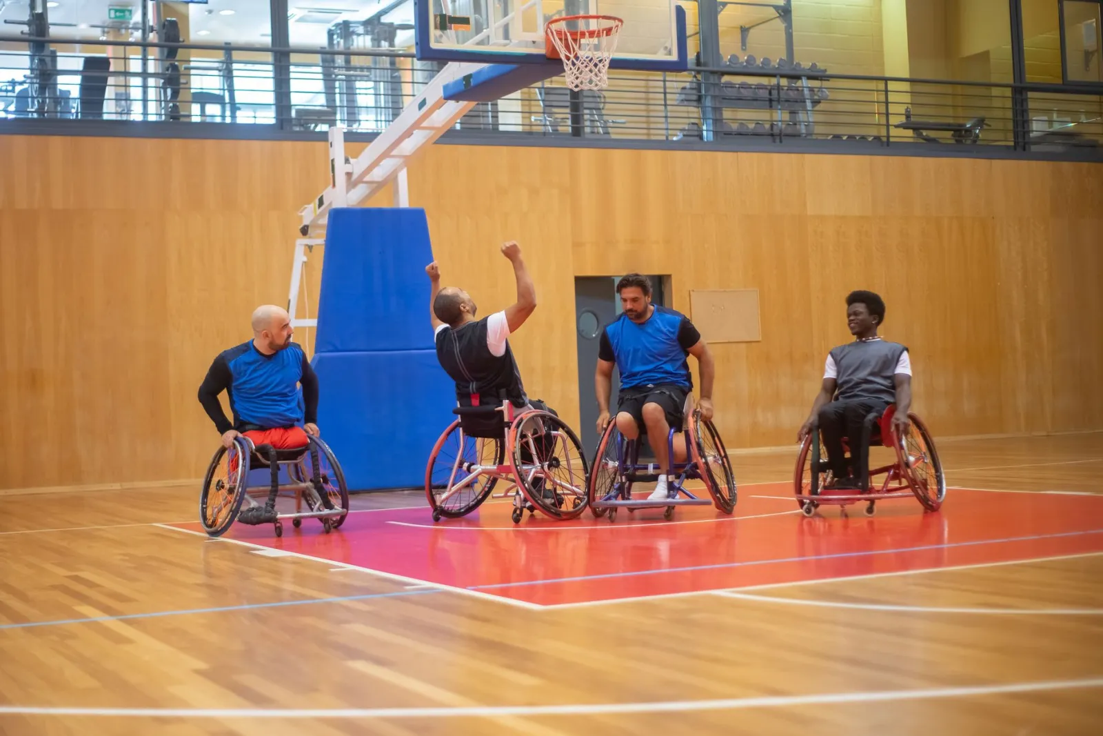 Four male wheelchair basketball players celebrating a successful shot on an indoor court.
