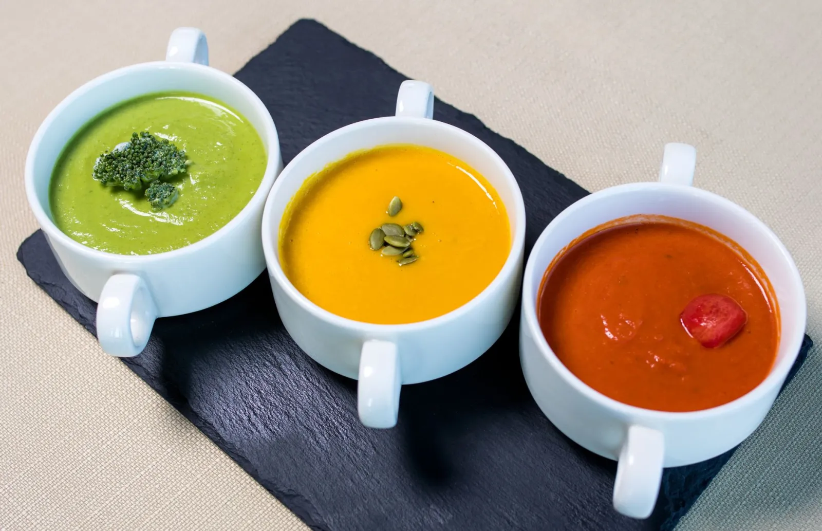 A variety of vegetarian soups presented in white mugs, featuring broccoli, pumpkin, and tomato flavors.